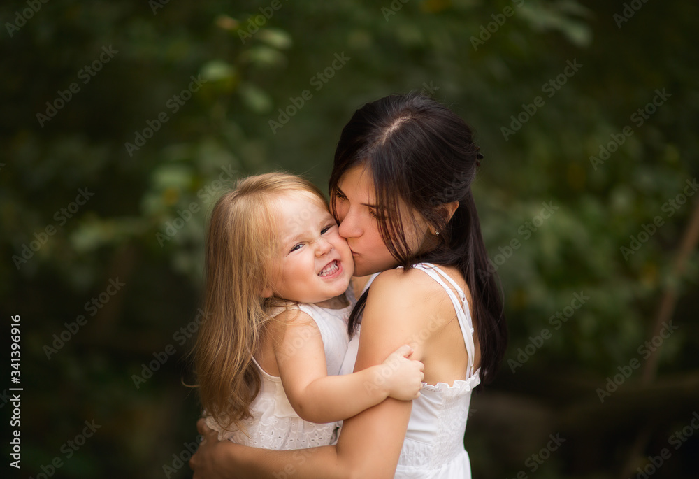 Young pretty black haired mother with blond little daughter in white dresses hugging outside in the summer