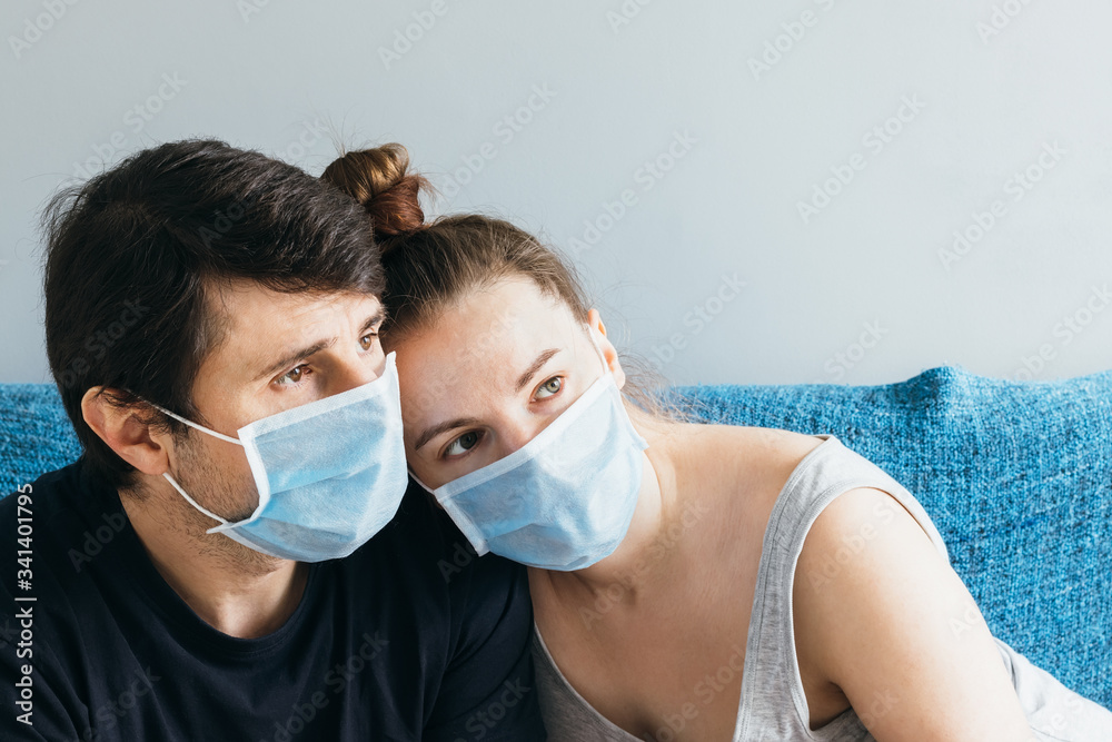 Married couple in blue medical face masks sitting on the couch, hugging and sadly looking into the distance. Man and woman, family of two, stay at home during quarantine