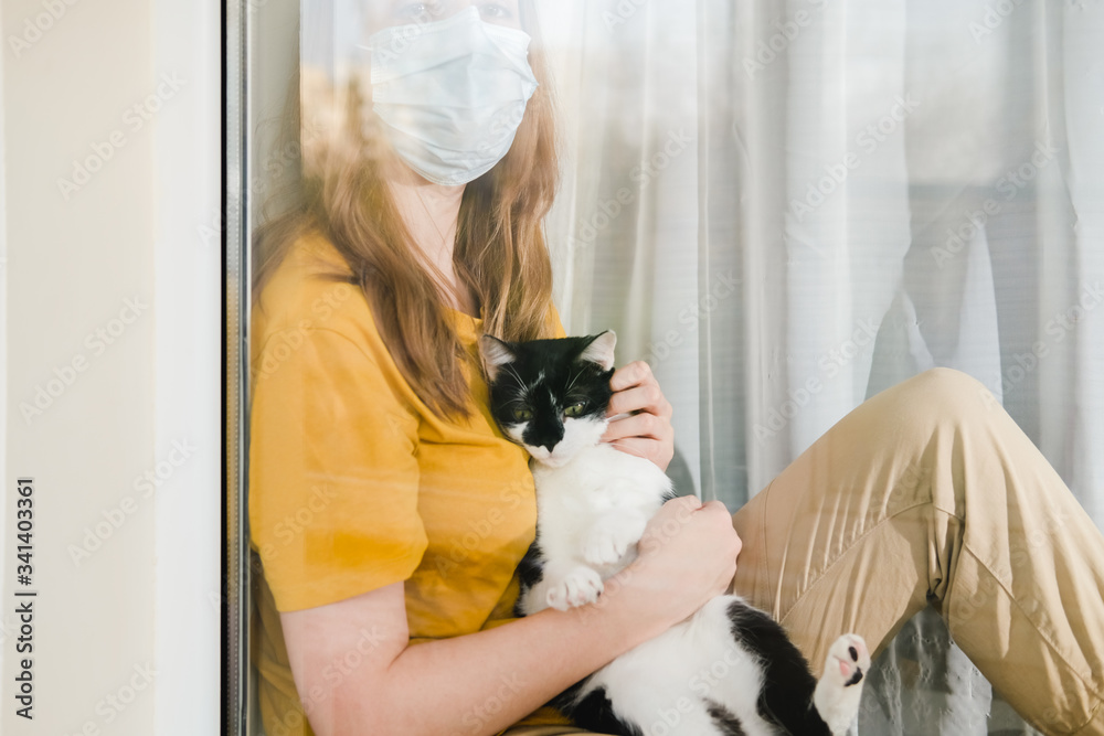 Sad girl in medical protective mask and yellow t-shirt sitting on the windowsill with a black and white Tuxedo cat during a pandemic quarantine coronavirus Covid-19. Isolation at home. Sad girl in medical protective mask and yellow t-shirt sitting on the windowsill with a black and white Tuxedo cat during a pandemic quarantine coronavirus Covid-19. Isolation at home.