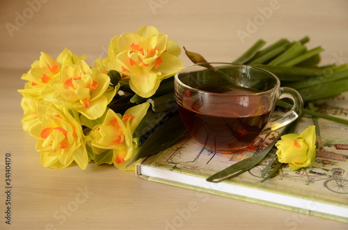 There is a glass cup of tea on the table, a notebook and a bouquet of beautiful yellow daffodils.