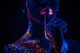 young sensual woman keep silence, show silence gesture at camera. cropped female with fluorescent prints on body, cosmic fantastic prints glowing in UV lights. body art concept