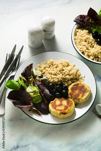 A simple vegan lunch or dinner for a family of two adults and a child. brown rice with soy okara and potato patties with fresh salad and olives on a marble table
