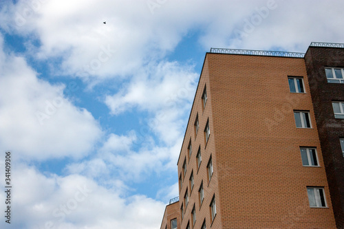 The wall of a high-rise building made of brick against the blue sky in the clouds. Corner view of a brick building against a blue sky. Windows on the facade of a high-rise building, side view.