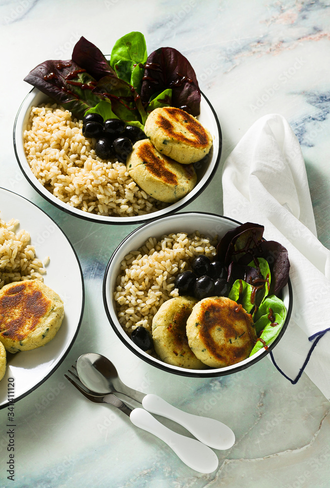 A simple vegan lunch or dinner for a family of two adults and a child. brown rice with soy okara and potato patties with fresh salad and olives on a marble table