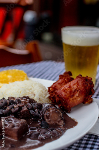 Brazilian feijoada on white plate. 
Accompanied by rice, sausage, orange and chopp beer.