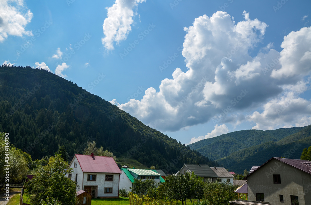 View on  Carpathian village Kolochava, Houses and coniferous foreston hills on background. Beautiful summer landscape, cloudy sky at bright sunny day. Travel background.