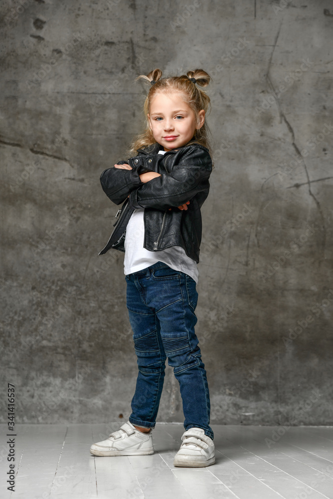Little cute smiling blond girl in stylish rock style black leather jacket, jeans and white sneakers standing in confident pose Little cute smiling blond girl in stylish rock style black leather jacket, jeans and white sneakers standing in confident pose