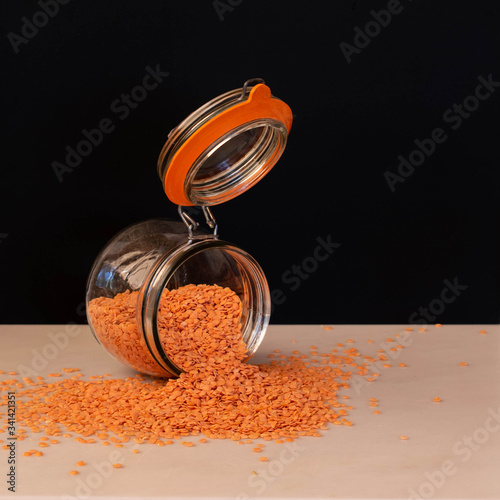 A glass kilner storage jar on its side with the lid wide open, red lentils, beans or pulses spill out on the a plain work surface.  The image is set against a plain black background. photo