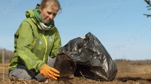 Close up woman eco volunteer picking trash a in black garbage bag on the forest glade. Early spring, bright blue sky, no leaves on the trees, windy weather. Earth day. Eco friendly concept