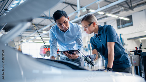 Instructor with a Tablet Computer is Giving a Task for a Future Mechanic. Female Student Inspects the Car Engine. Assistant is Checking the Cause of a Breakdown in the Vehicle in a Car Service.  © Gorodenkoff