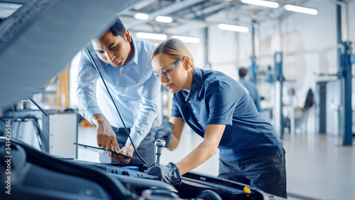 Instructor with a Tablet Computer is Giving a Task for a Future Mechanic. Female Student Inspects Car Engine and Uses Ratchet. Assistant is Checking Cause of Breakdown in Vehicle in a Car Service. 
