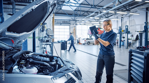 Female Mechanic Uses a Tablet Computer with an Augmented Reality Diagnostics Software. Specialist Inspecting the Car in Order to Find Broken Components Inside the Engine Bay. Modern Car Service.