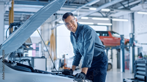 Handsome Professional Car Mechanic is Working on a Vehicle in a Service. Repairman Looks Happy While Using a Ratchet. Specialist is Wearing Safety Glasses. He Looks at a Camera and Smiles.