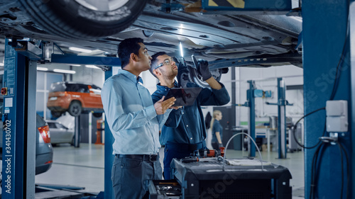 Manager Checks Data on a Tablet Computer and Explains the Breakdown to a Mechanic. Car Service Employees Inspect the Bottom of the Car with a LED Lamp. Modern Clean Workshop.