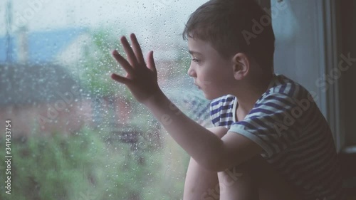 a little boy sits outside a rainy window and is sad. A window in rainy weather behind which a small sad boy sits, a child is hard going through self-isolation, orphaned children in orphanages and boar
