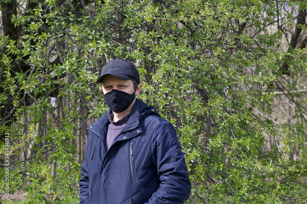 Portrait of a young man walking in a spring park. On his face is a black cloth mask. Dressed in a spring jacket, cap on his head. A walk in the fresh air during a pandemic.