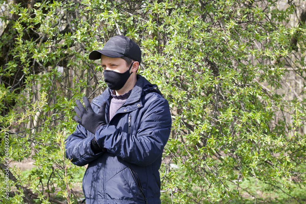A young man walks through a spring park and gestures. On his face is a black cloth mask. Dressed in a spring jacket, cap on his head. A walk in the fresh air during a pandemic.