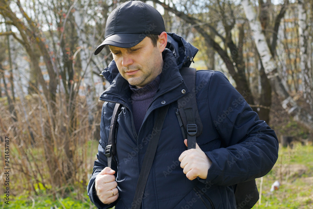A young man walks in the spring park. Corrects a backpack over his shoulders. Dressed in a spring jacket, cap on his head. Walk in the fresh air.