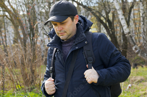 A young man walks in the spring park. Corrects a backpack over his shoulders. Dressed in a spring jacket, cap on his head. Walk in the fresh air.