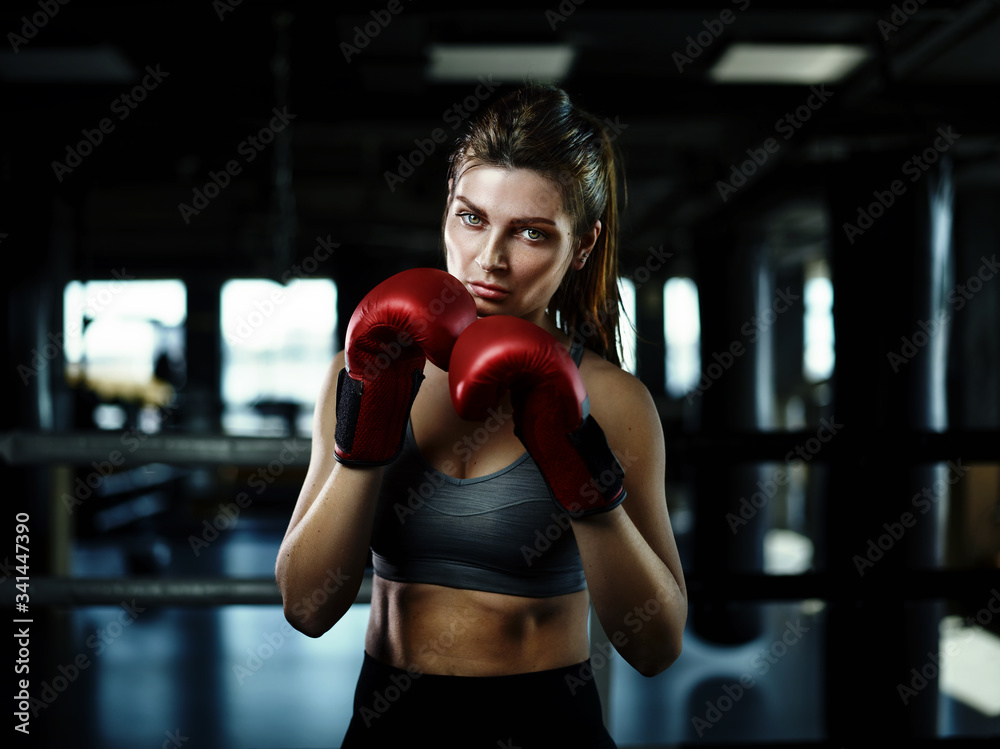 Portrait of attracive female boxer in red gloves looking confidently at camera while posing in dark ring