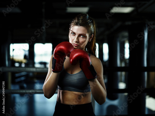 Portrait of attracive female boxer in red gloves looking confidently at camera while posing in dark ring © Comeback Images