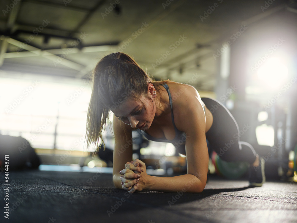 Fototapeta premium Backlit shot of determined tired young woman doing forearm plank exercise during workout in gym