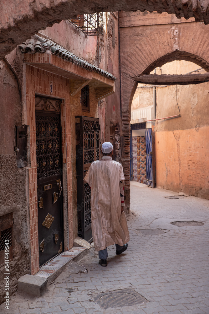 Muslim man with djellaba and kufi strolling alley with shop on the street, blurred background