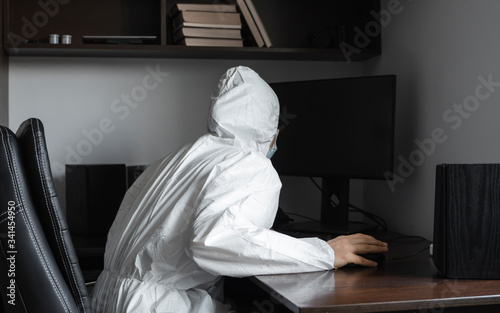 Man in medical mask and protective suit, rubber gloves sits at home and works with pc at the table during quarantine. Designer, artist, architect, businessman at remote work in a pandemic covid. © Volodymyr