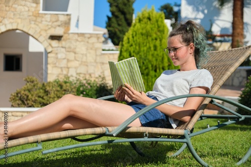 Teenager girl in glasses reading book, resting on sunbed standing in backyard