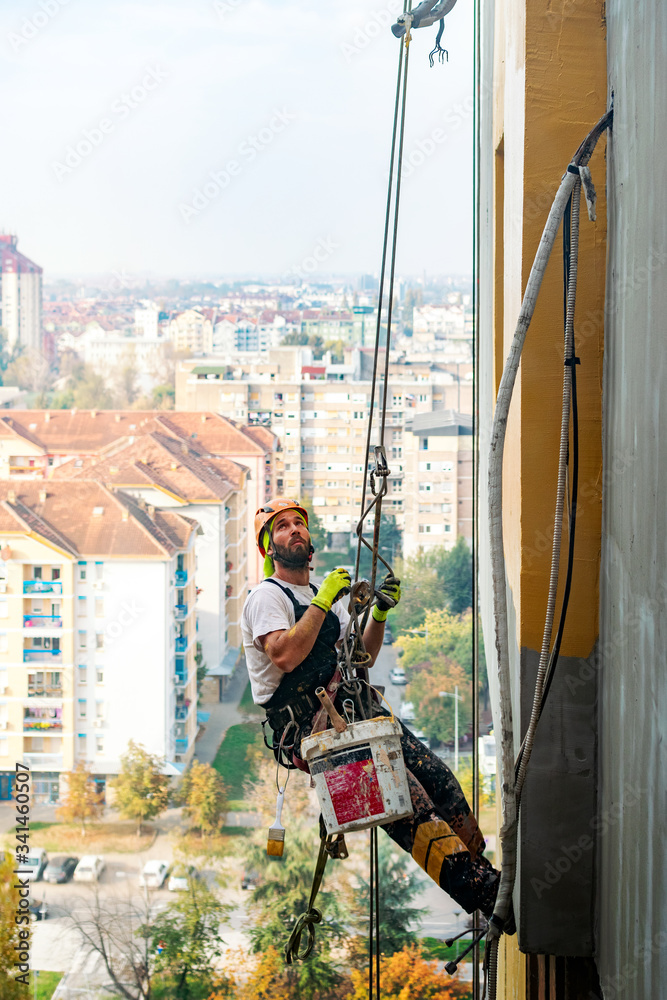 Industrial climber hanging on the rope while painting the exterior facade wall of the tall apartment building. Industrial alpinism and high risk work scene concept image