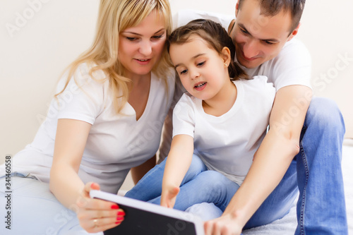 An attractive happy, family of mother, father and daughter sitting on a sofa at home having fun using a tablet computer