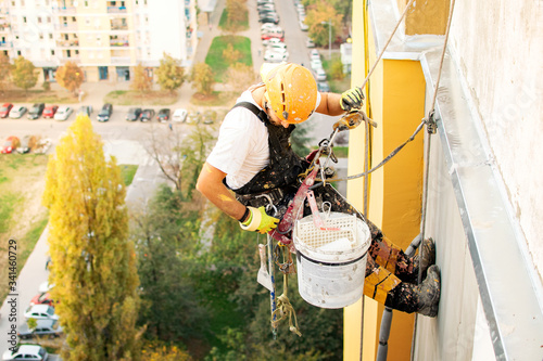 Industrial climber hanging on the rope while painting the exterior facade wall of the tall apartment building. Industrial alpinism and high risk work scene concept image