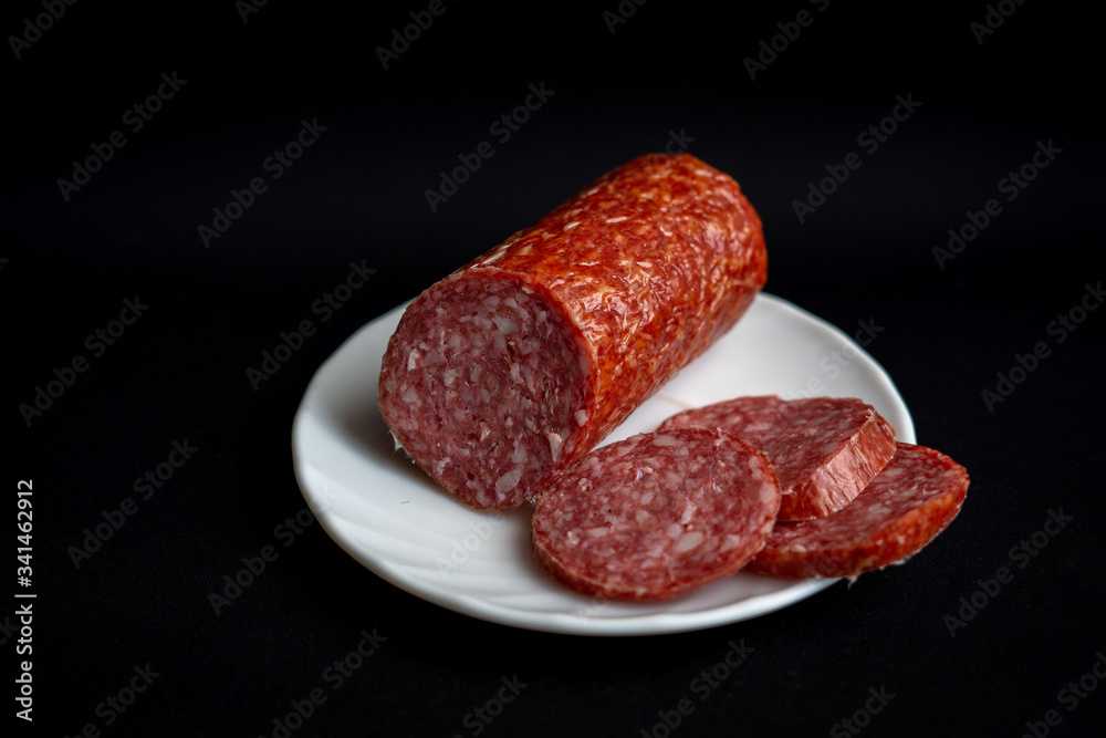 Slices of tasty delicious pepperoni sausage on white plate on dark black background Slices of tasty delicious pepperoni sausage on white plate on dark black background