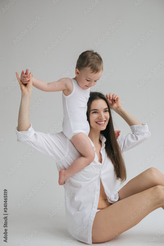 Young beautiful brunette mom plays with her little baby son. Stylishly dressed in white clothes. Hug, happy together, play. Studio portrait. Motherhood concept, tender feelings. Young beautiful brunette mom plays with her little baby son. Stylishly dressed in white clothes. Hug, happy together, play. Studio portrait. Motherhood concept, tender feelings.