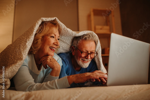 Close photo of lovely elderly couple browsing internet together on laptop in bed covered with blanket.
