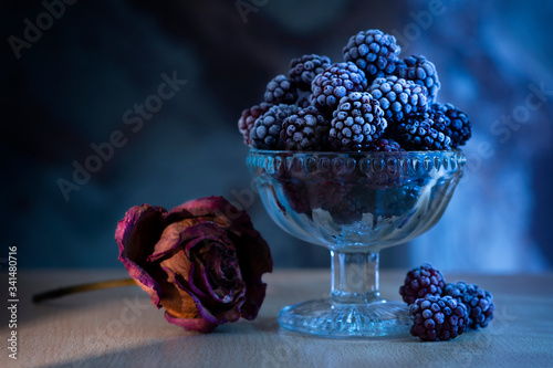 Frozen blackberry in a vase in the moonlight on a table, next to a dried rose on a table on which yellow light falls from an ajar door