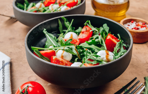 Healthy arugula salad with mozzarella cheese and cherry tomatoes in black bowls on beige rustic background.