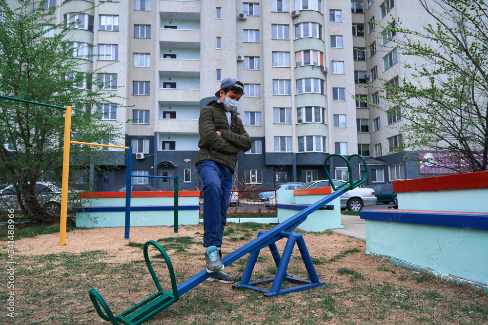 teen boy stands by seesaw on playground near high-rise buildings with apartments, a medical mask on his face protects against viruses and dust