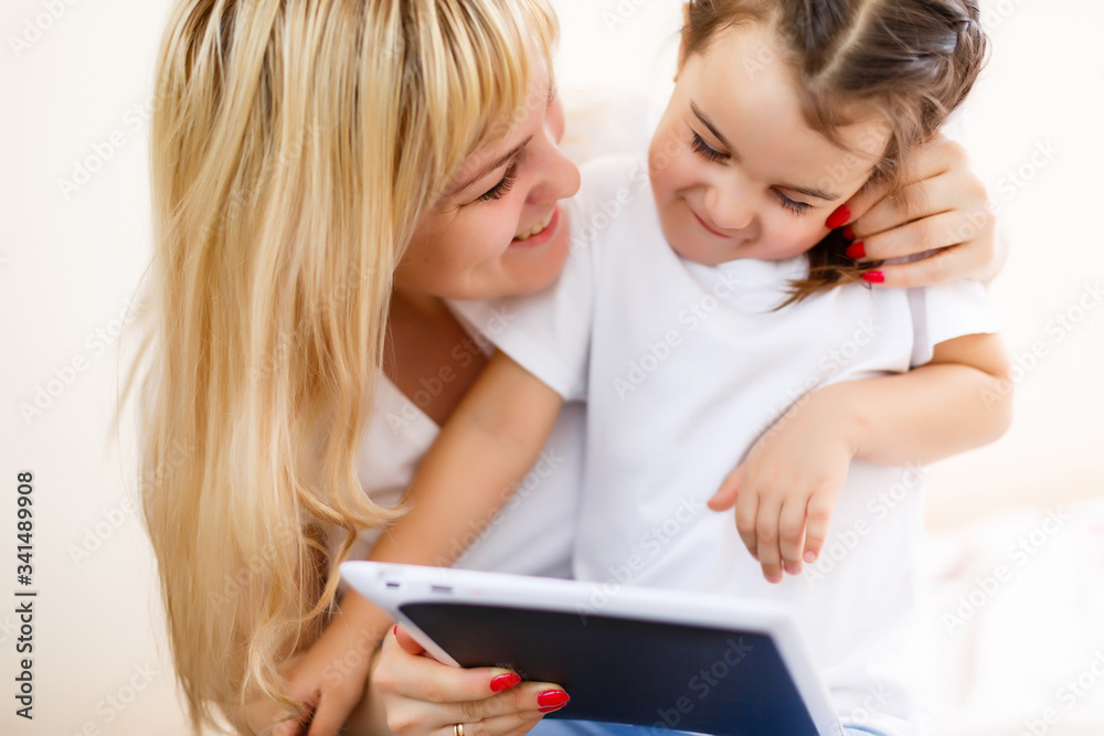 Happy family mom and kid daughter using digital tablet sitting on sofa, smiling parent mother with child girl holding tablet looking at screen do online shopping make video call watch cartoons Happy family mom and kid daughter using digital tablet sitting on sofa, smiling parent mother with child girl holding tablet looking at screen do online shopping make video call watch cartoons