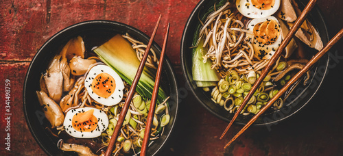 Flat-lay of traditional japanese Ramen bowls with chicken meat and shiitake mushrooms and bamboo chopsticks over dark red damaged wooden table background, top view, wide composition