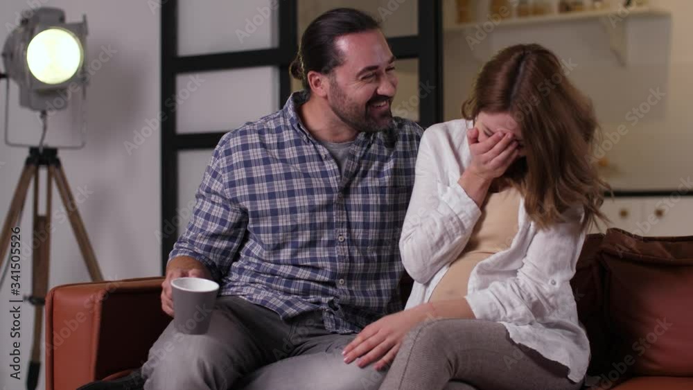 Close-up of laughing future parents chatting fun while sitting on sofa in domestic room. Happy husband and pregnant wife enjoying conversation while spending free time together at home