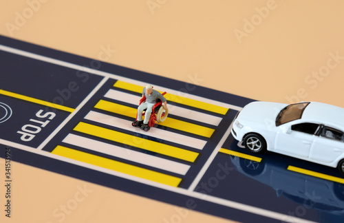A miniature man in a wheelchair crossing a crosswalk and a miniature car standing on a stop line. 