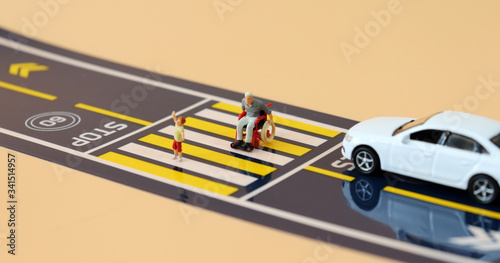 A miniature child with a hand raised and a miniature man in a wheelchair crossing a crosswalk. 