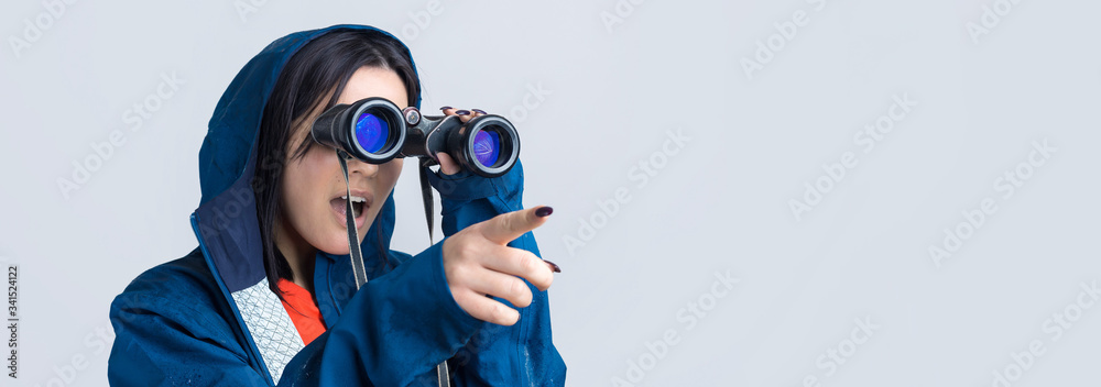Φωτογραφία A tourist girl in a blue raincoat holds binoculars in her hands and looks into the distance, spies