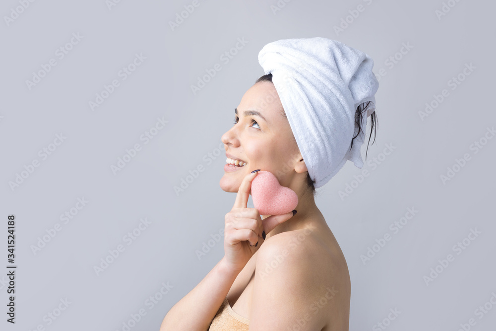 Beauty portrait of woman in white towel on head with a sponge for a body in view of a pink heart. Skincare cleansing eco organic cosmetic spa relax concept. Beauty portrait of woman in white towel on head with a sponge for a body in view of a pink heart. Skincare cleansing eco organic cosmetic spa relax concept.