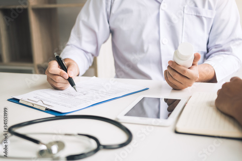 Confident doctor man holding a pill bottle and writing while talking with senior patient and reviewing his medication at office room.