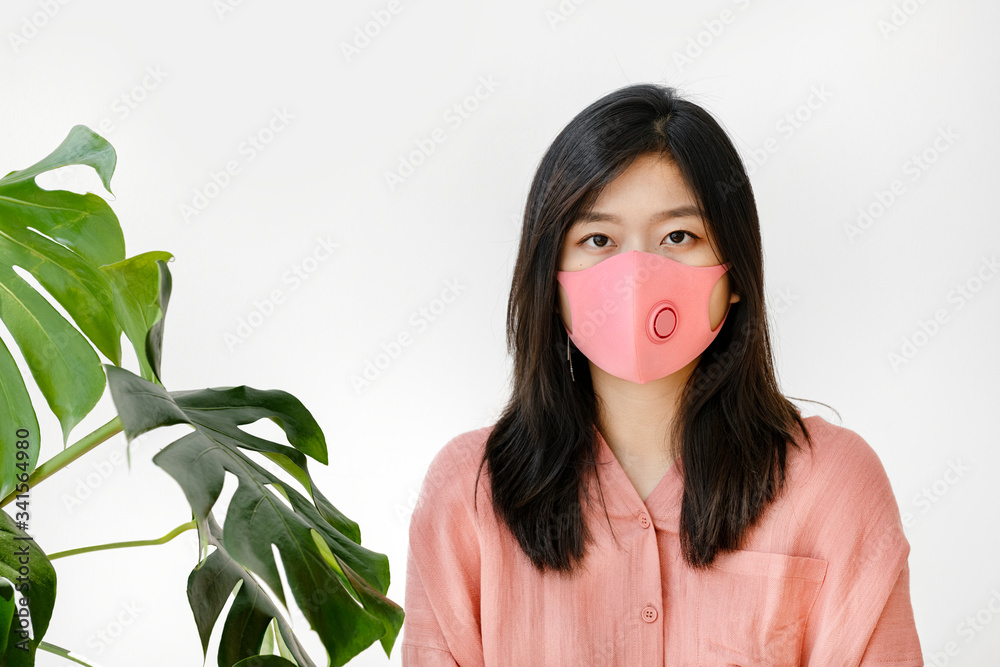 Asian woman wearing a mask standing next to a monstera plant Asian woman wearing a mask standing next to a monstera plant