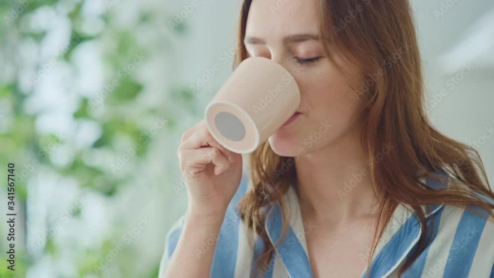 Authentic Portrait of a Beautiful Young Female in a Bright Room at Home Drinking Tea or Coffee from a Cup. Attractive Brunette Woman Poses for the Camera and Gently Smiles. Happy Morning Routine. Authentic Portrait of a Beautiful Young Female in a Bright Room at Home Drinking Tea or Coffee from a Cup. Attractive Brunette Woman Poses for the Camera and Gently Smiles. Happy Morning Routine.