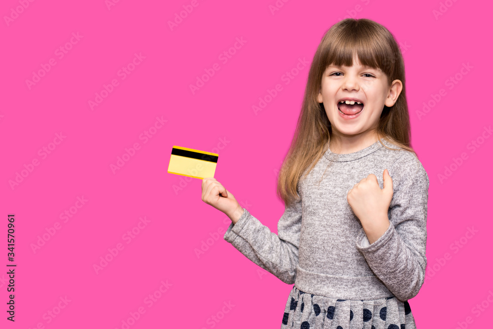 Smiling Pretty child girl with hands up in winner pose holding credit card while rejoices over purple background Smiling Pretty child girl with hands up in winner pose holding credit card while rejoices over purple background
