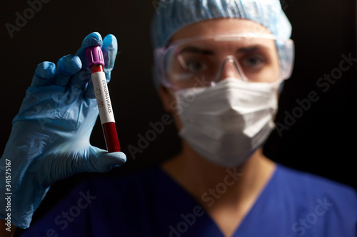 medicine, health and pandemic concept - close up of young female doctor or nurse wearing goggles, glove and face protective mask holding beaker with coronavirus blood test over black background